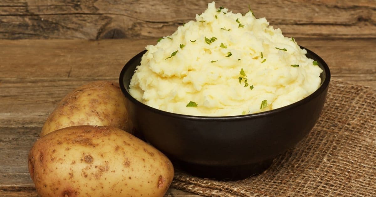 A black ceramic bowl of mashed potatoes on a wooden table, next to whole unpeeled raw potatoes